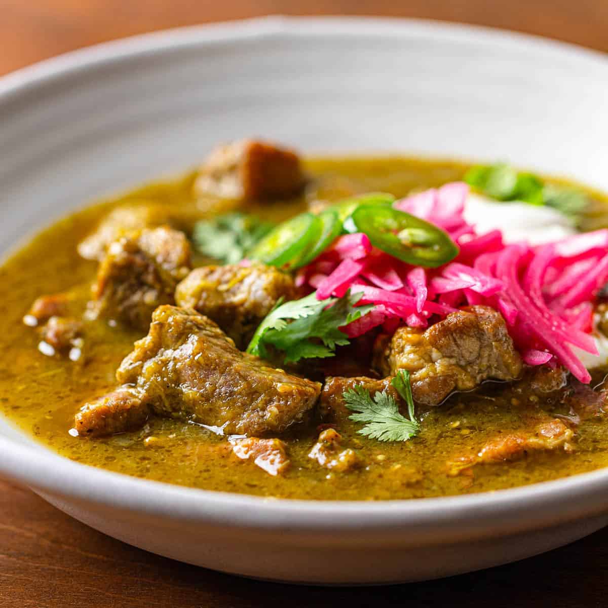White bowl of pork chili verde garnished with sour cream and pickled onions.