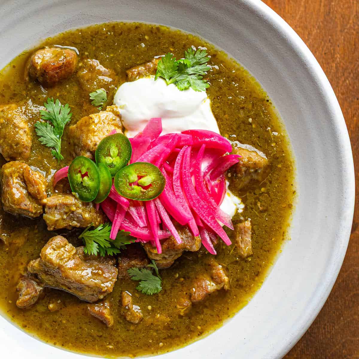 White bowl of pork chili verde with creamy and pickled toppings.