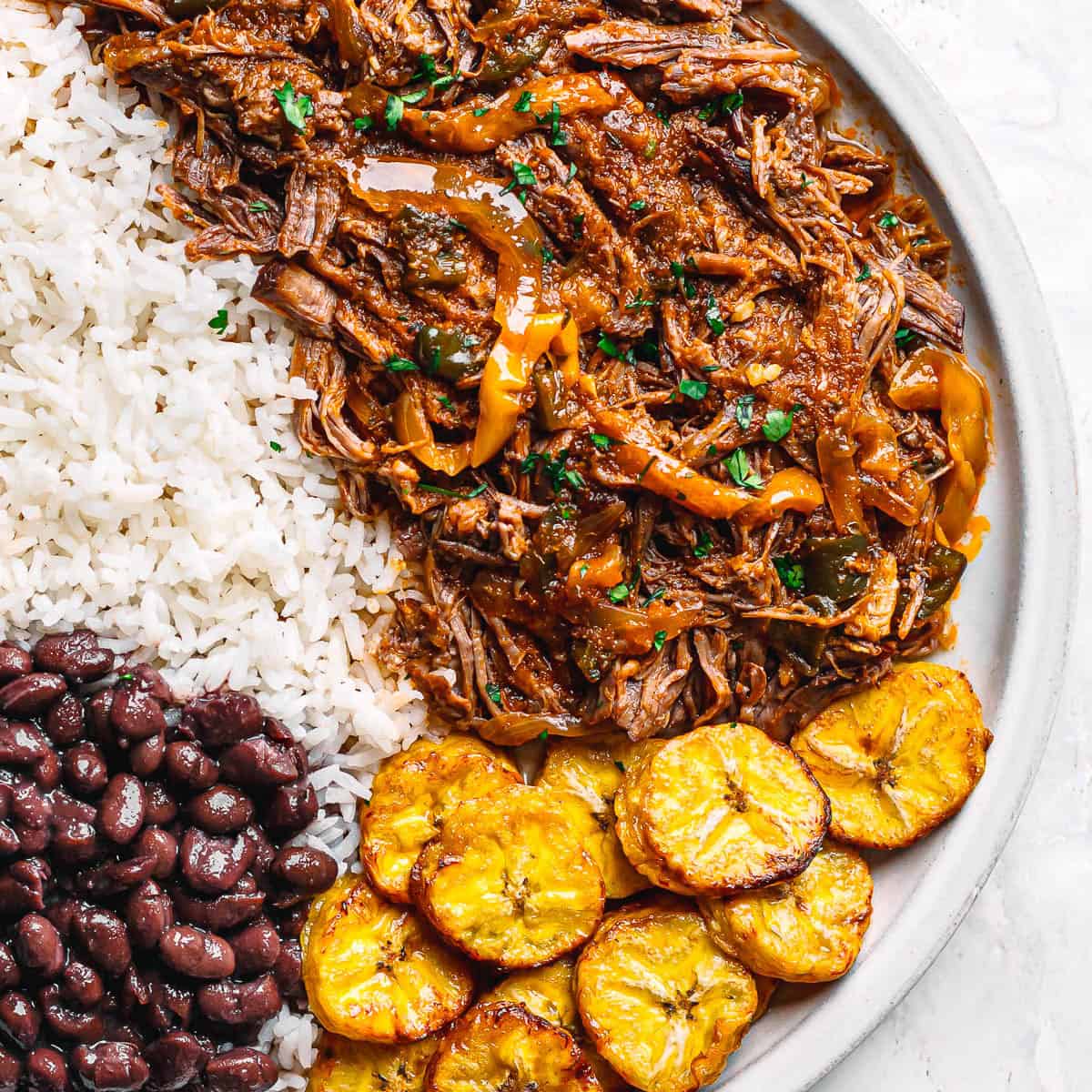 Authentic ropa vieja with rice, beans, and plantains on a white plate.