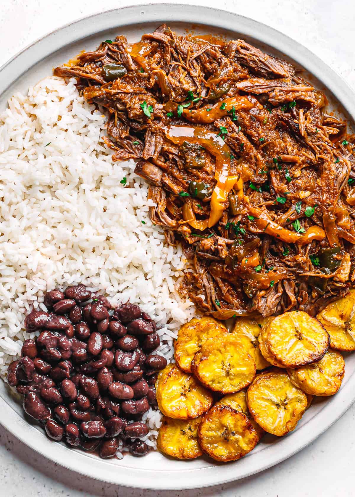 Close-up of ropa vieja with rice, beans, and plantains on a dinner plate.
