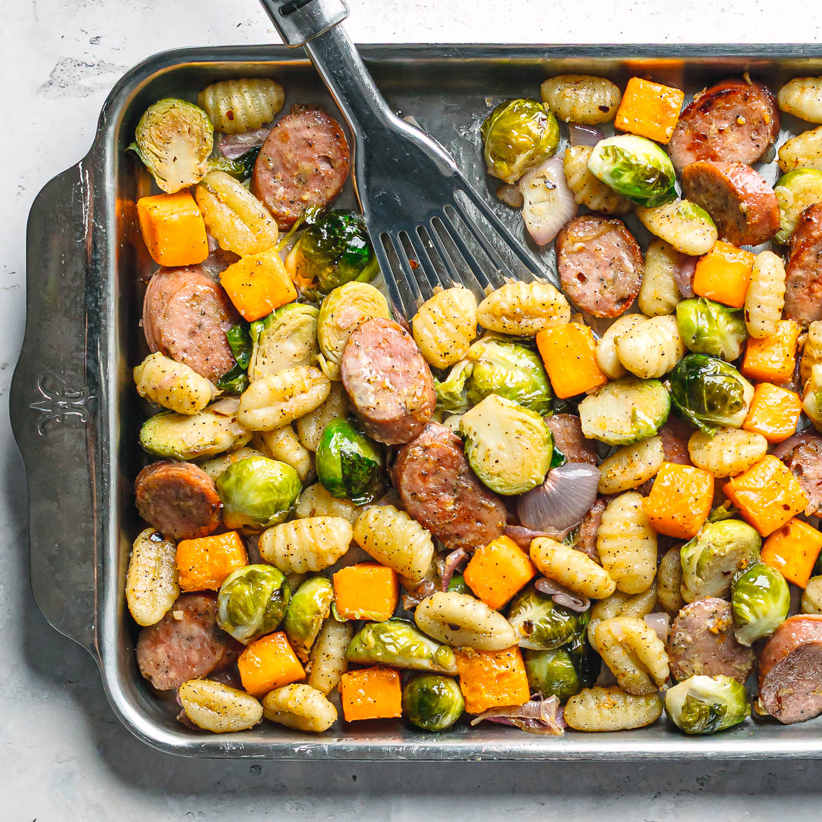 Sheet pan chicken sausage with Brussels sprouts, butternut squash, gnocchi, and shallots roasted to golden brown in a stainless steel sheet pan.