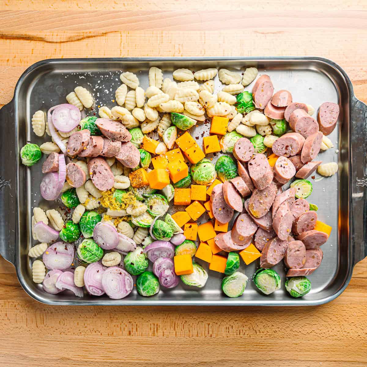 Brussels sprouts, squash, shallots, gnocchi, and sliced chicken sausage spread on an extra large stainless steel sheet pan.
