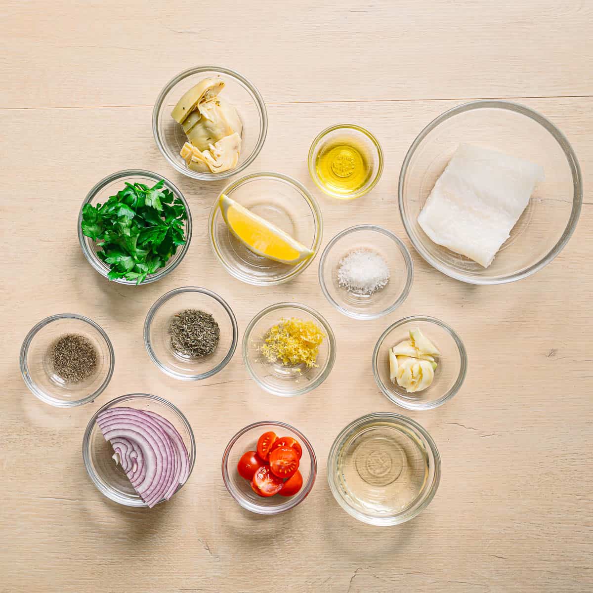 Ingredients for Mediterranean baked cod including cod fillets, cherry tomatoes, artichoke hearts, garlic, lemon, olive oil, and fresh herbs.