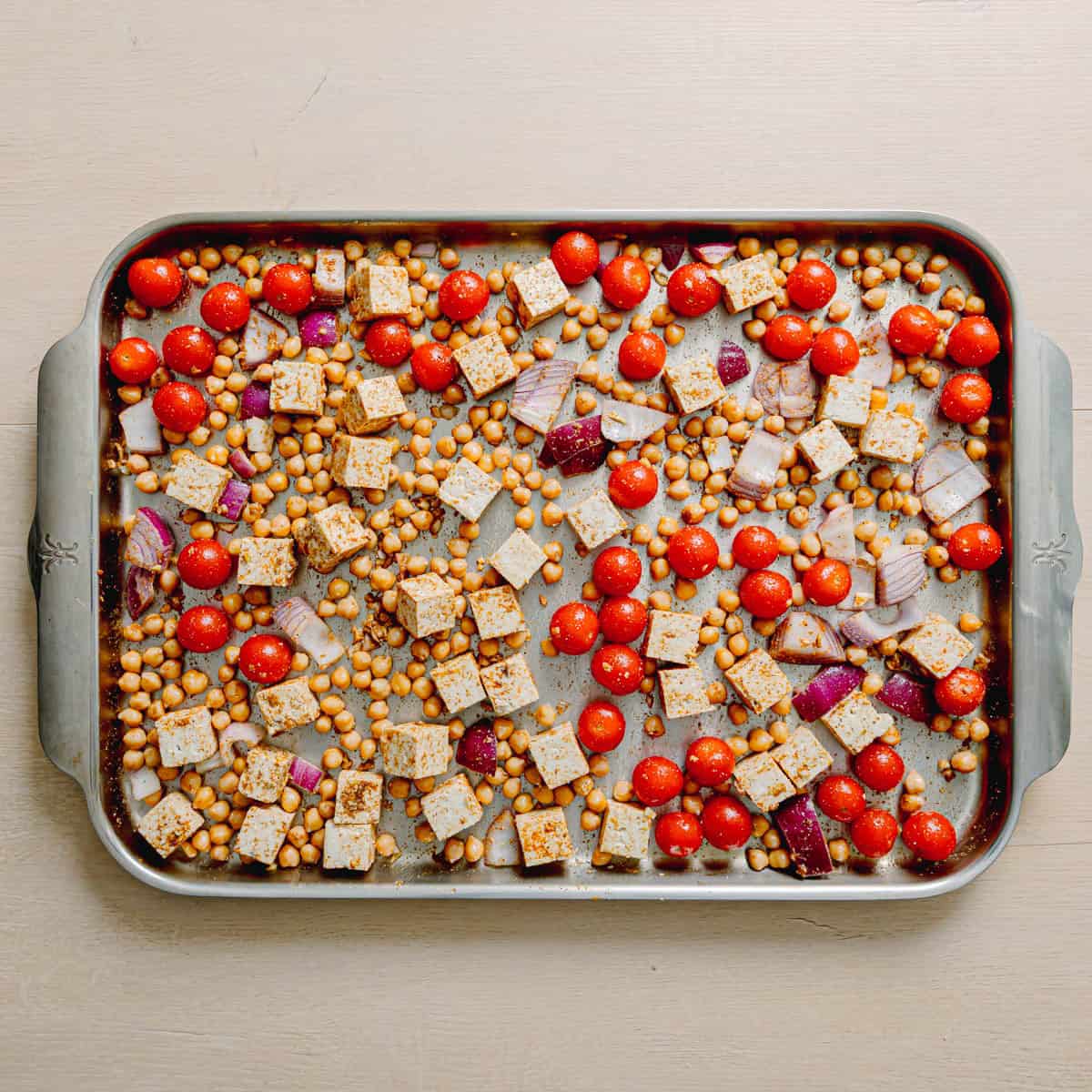 Mixing chickpeas tofu and tomatoes with masala and Indian spices on a sheet pan.