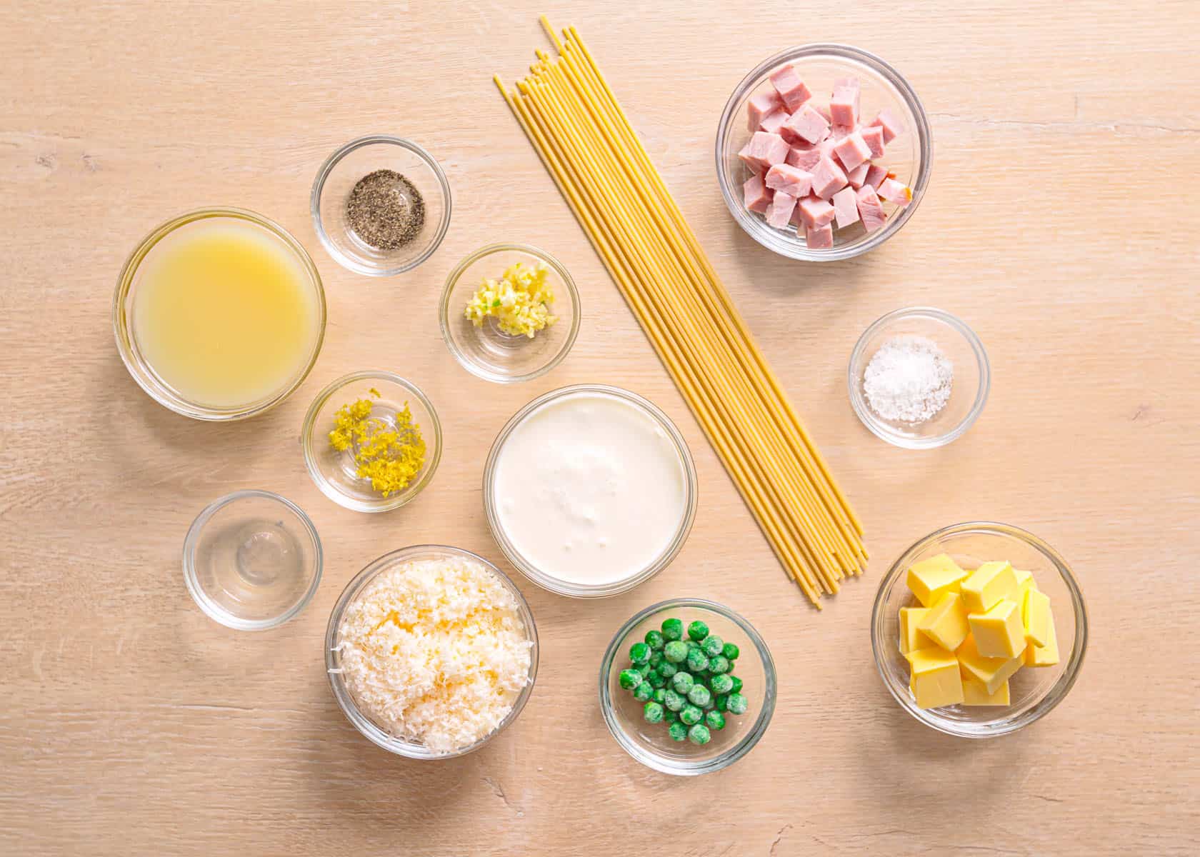 Ingredients for pasta with ham and peas including spaghetti, garlic, heavy cream, lemon, broth, and parmesan cheese arranged on a kitchen counter.