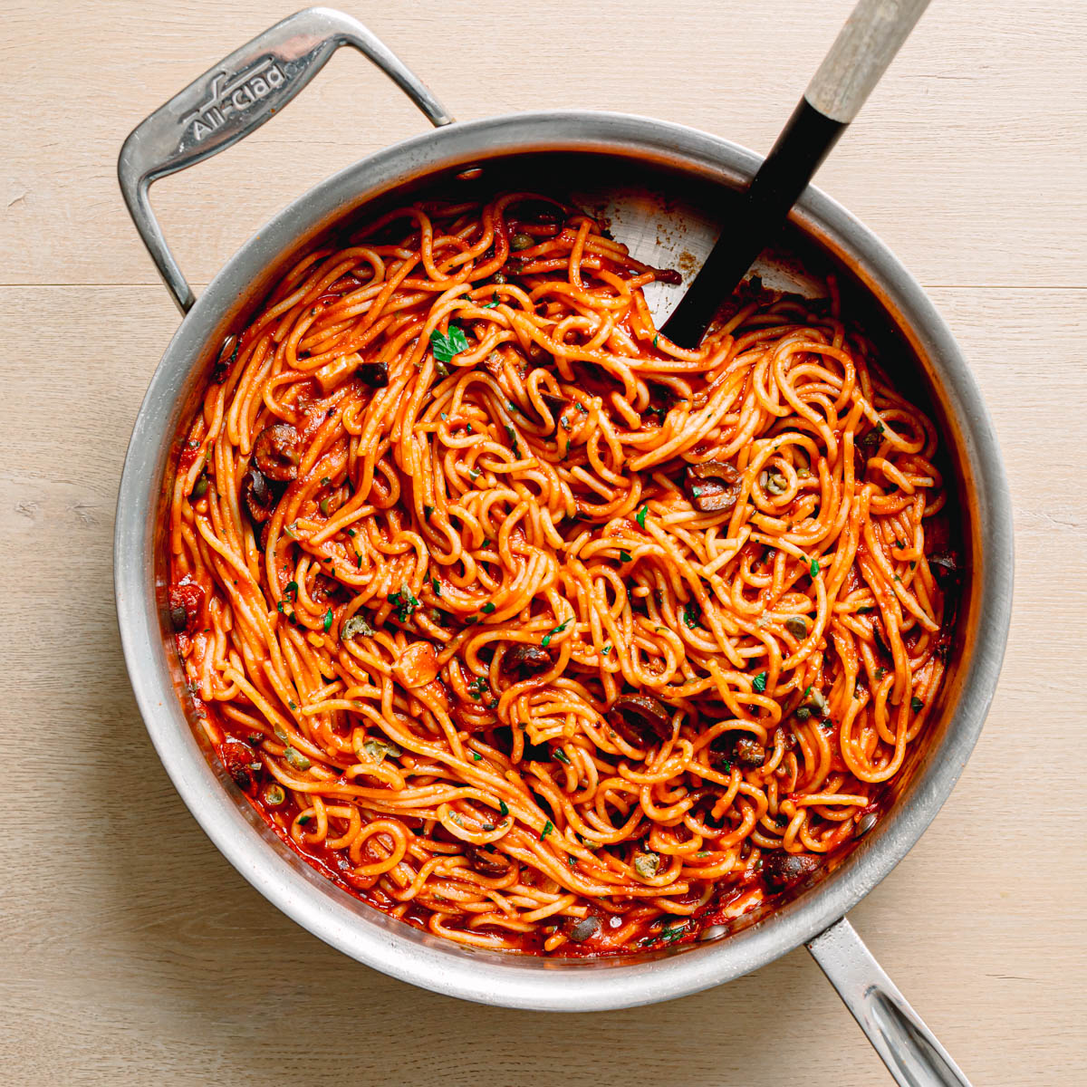 Overhead view of pasta puttanesca recipe in a large skillet garnished with fresh parsley and olives.