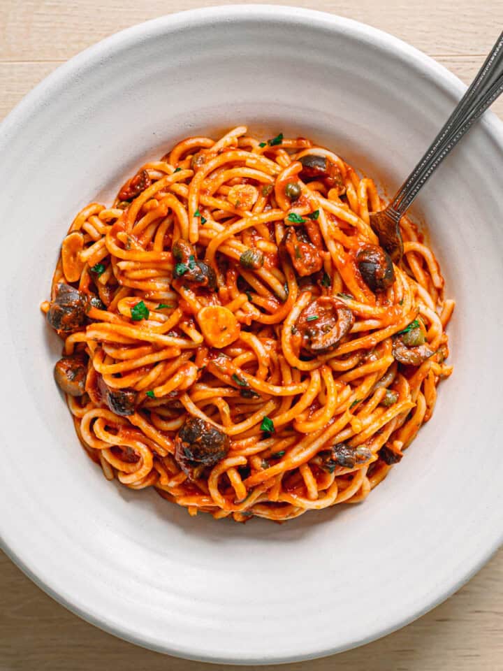 Close up of easy pasta puttanesca recipe served in a white ceramic bowl with spaghetti, olives, capers and fresh parsley.