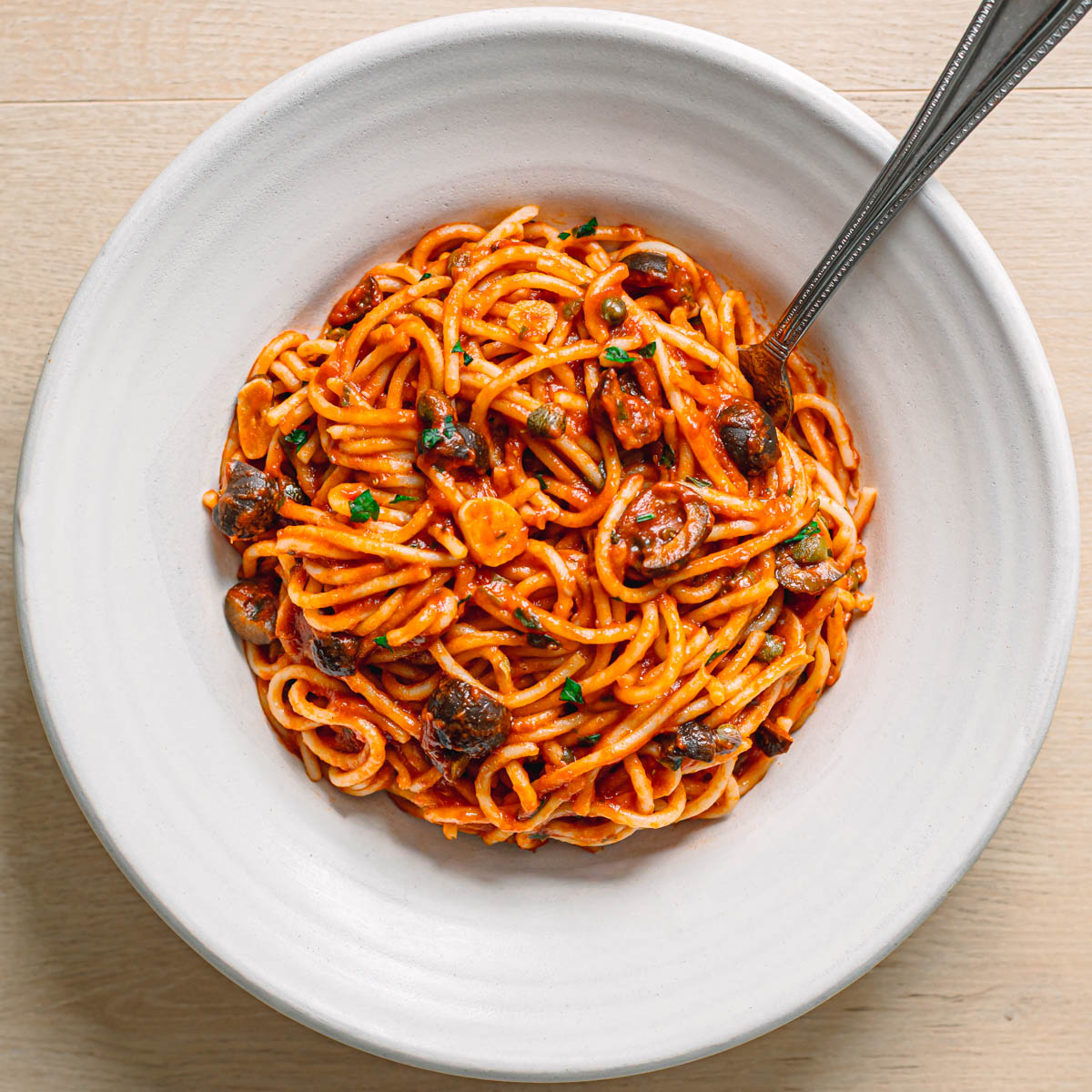 Close up of easy pasta puttanesca recipe served in a white ceramic bowl with spaghetti, olives, capers and fresh parsley.