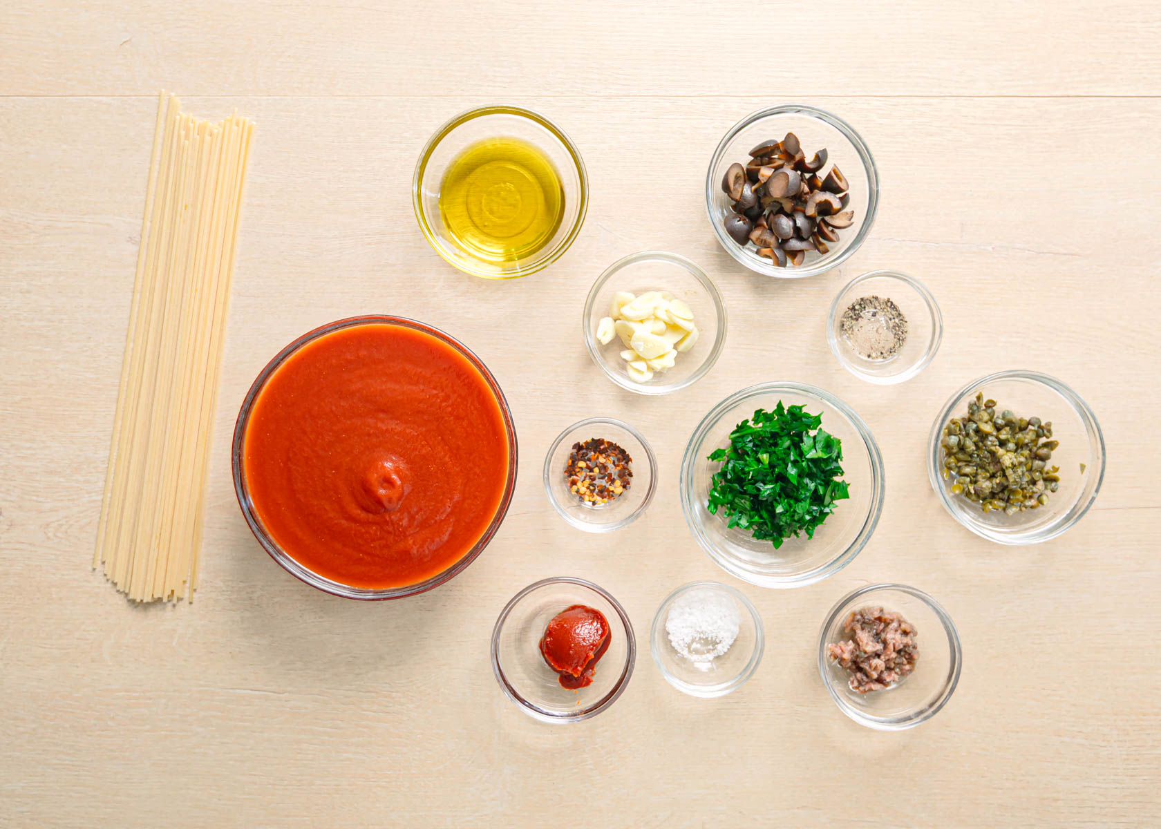 Pasta puttanesca recipe ingredients laid out on a table including spaghetti, olives, capers, anchovy fillets, garlic, crushed tomatoes, tomato paste, parsley, olive oil, red pepper flakes, salt and black pepper.