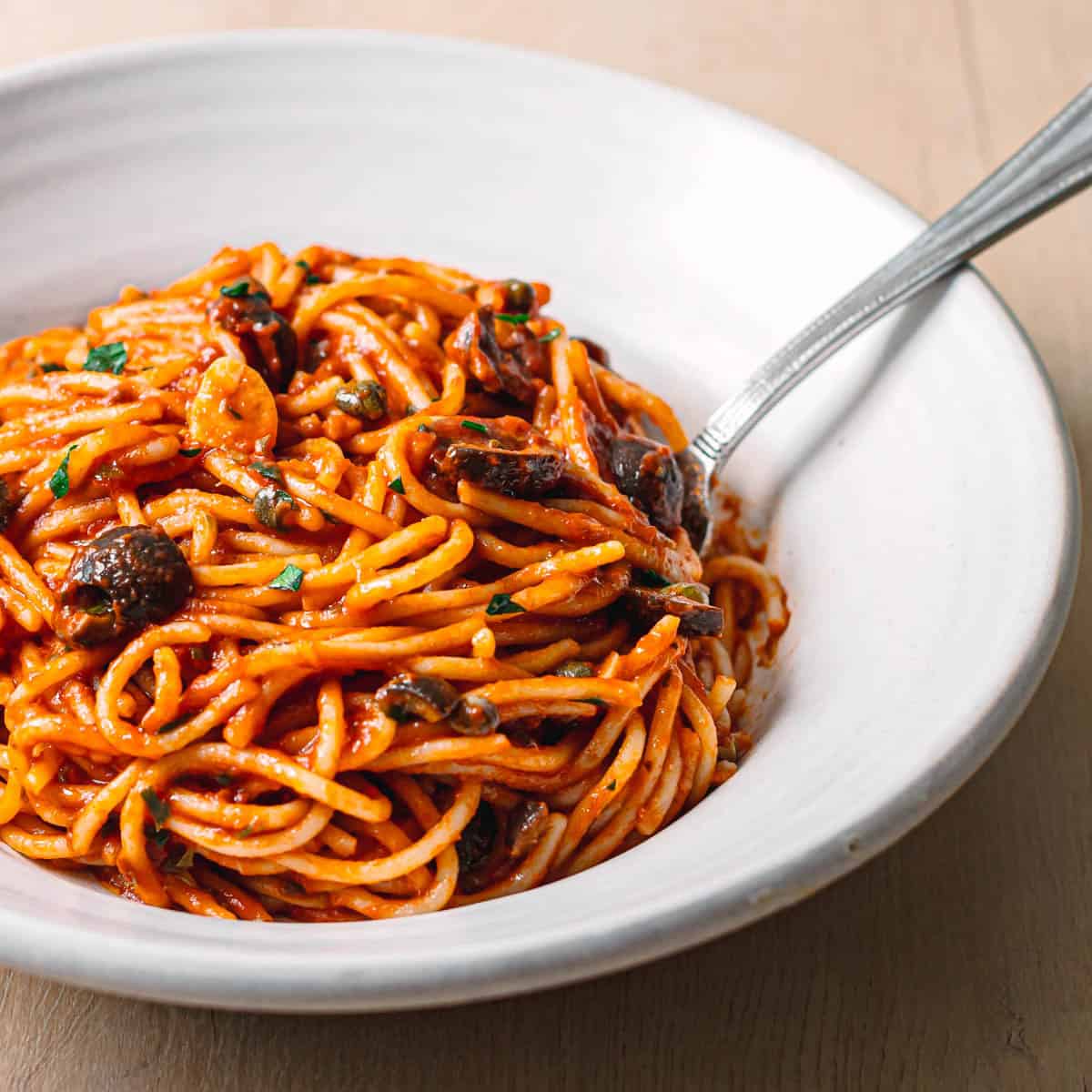 Side angle of pasta puttanesca recipe in a ceramic bowl garnished with fresh parsley, red pepper flakes and a drizzle of olive oil.