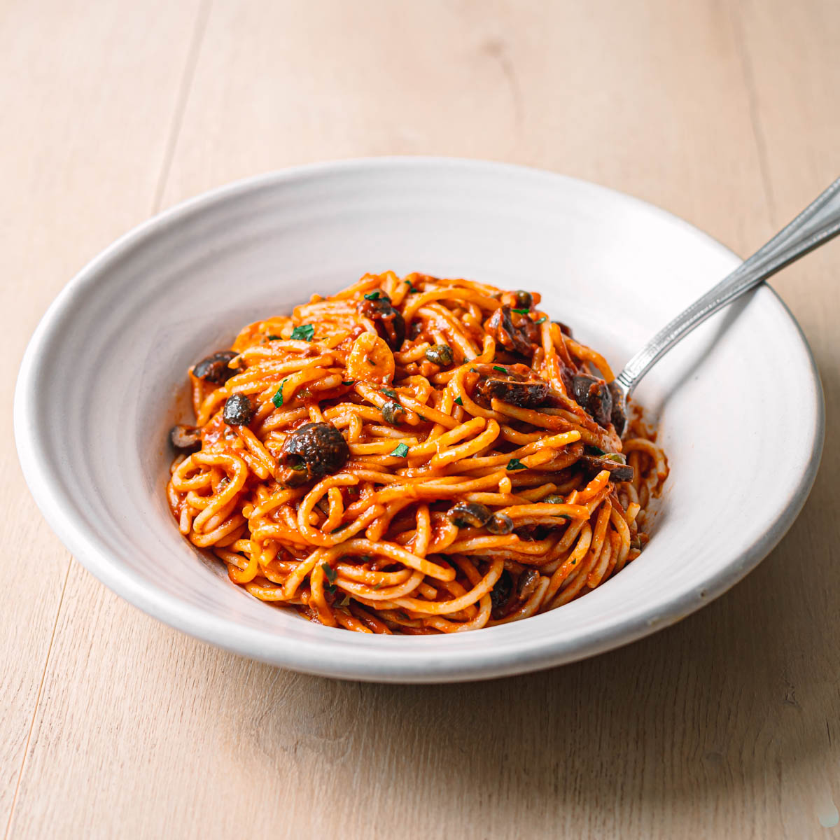 Side angle of homemade pasta puttanesca recipe in a ceramic bowl in natural light showing spaghetti coated in rich tomato sauce with olives capers and parsley.
