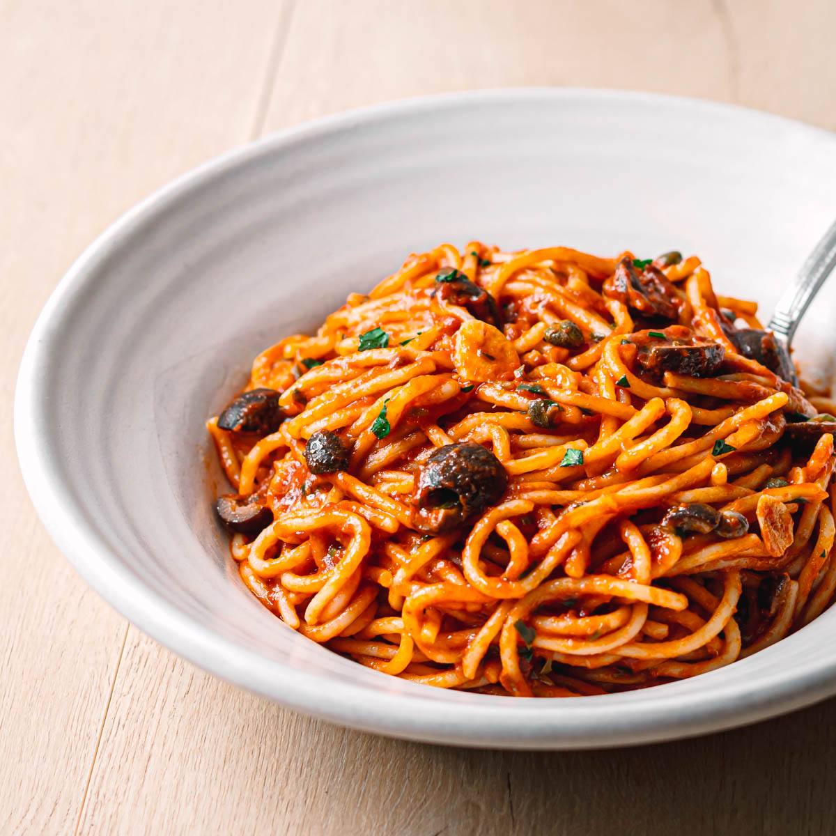Side angle view of pasta puttanesca recipe in a ceramic bowl showing rich tomato sauce with spaghetti olives and capers.