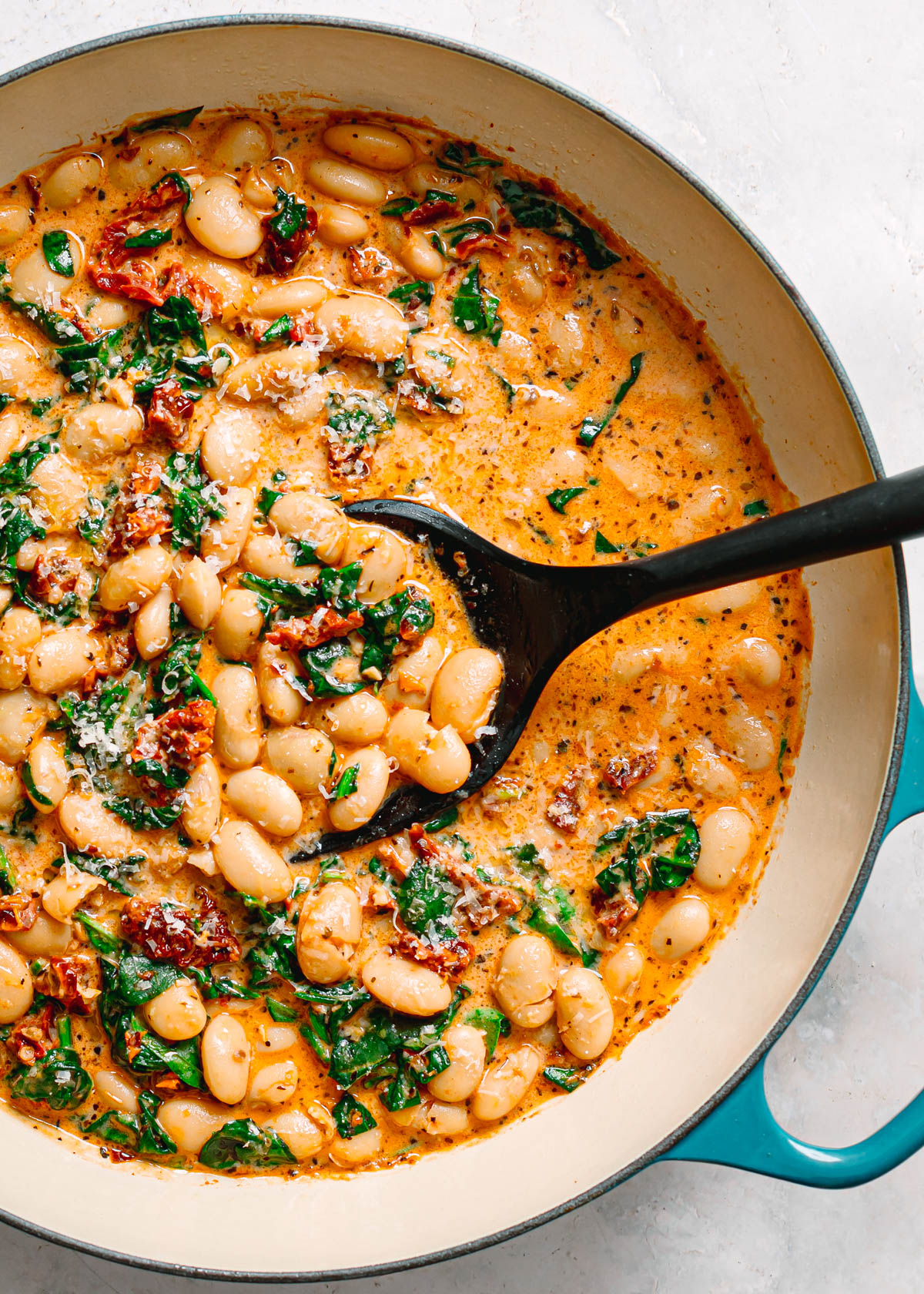 Close up overhead shot of a spoon scooping creamy marry me butter beans with spinach and sun dried tomatoes in a teal Le Creuset pot.