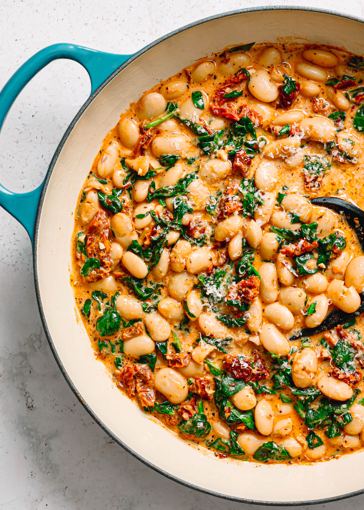 Overhead close up of creamy marry me butter beans with spinach, sun dried tomatoes, and parmesan in a teal Le Creuset pot.